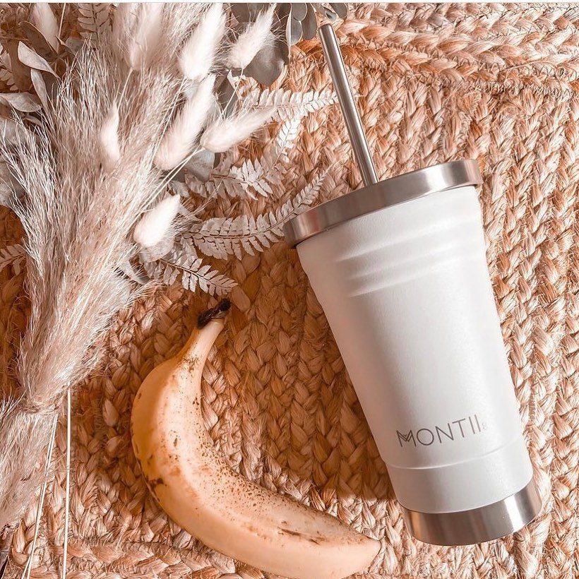 White insulated tumbler with straw, a banana, and dried grass on woven mat