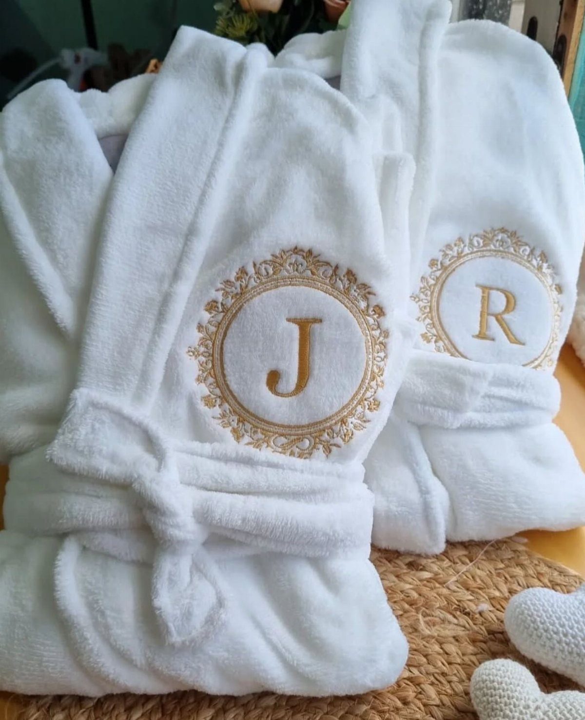 Two plush white bathrobes with gold embroidered initials on a woven mat.