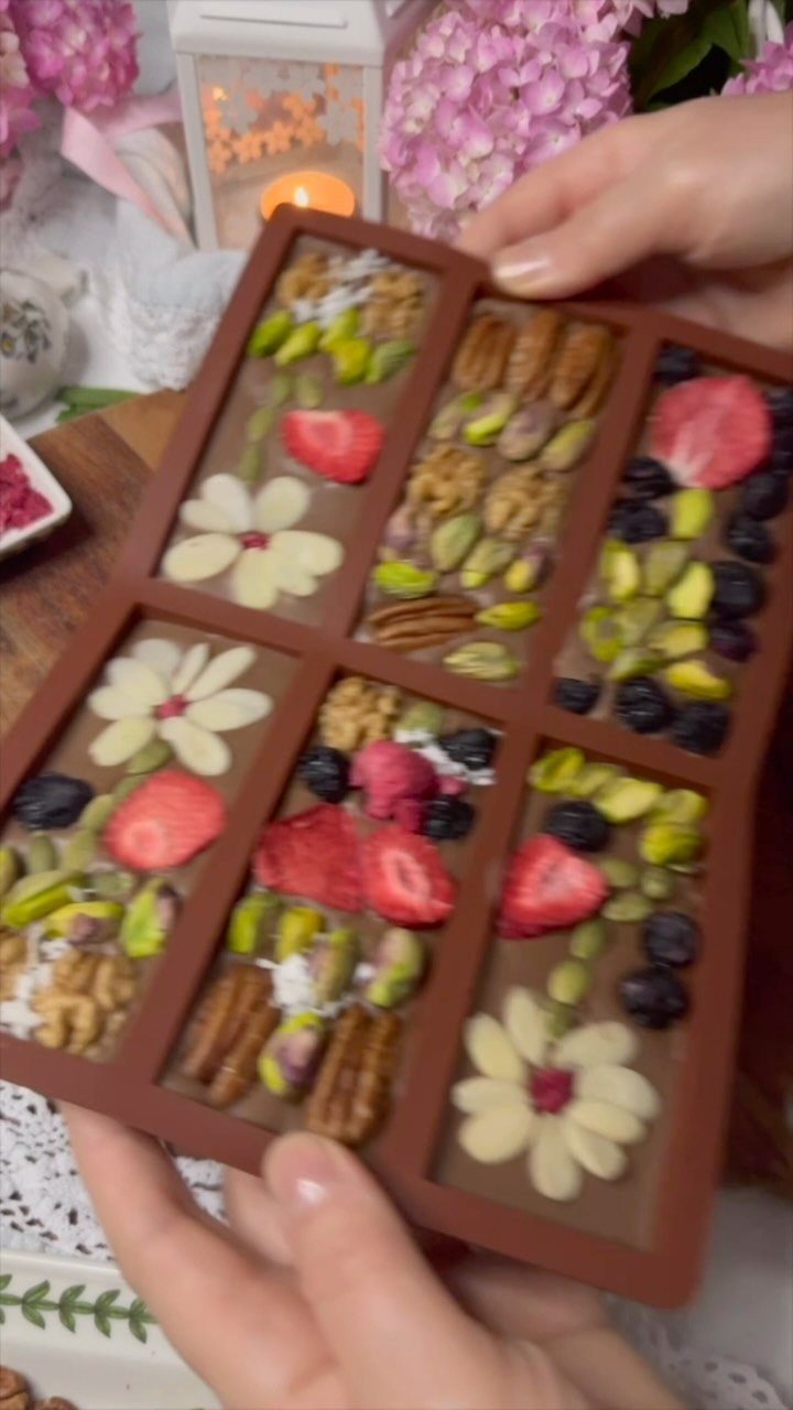 Decorated chocolate bar with dried fruits, nuts, and white chocolate flowers held by hands.