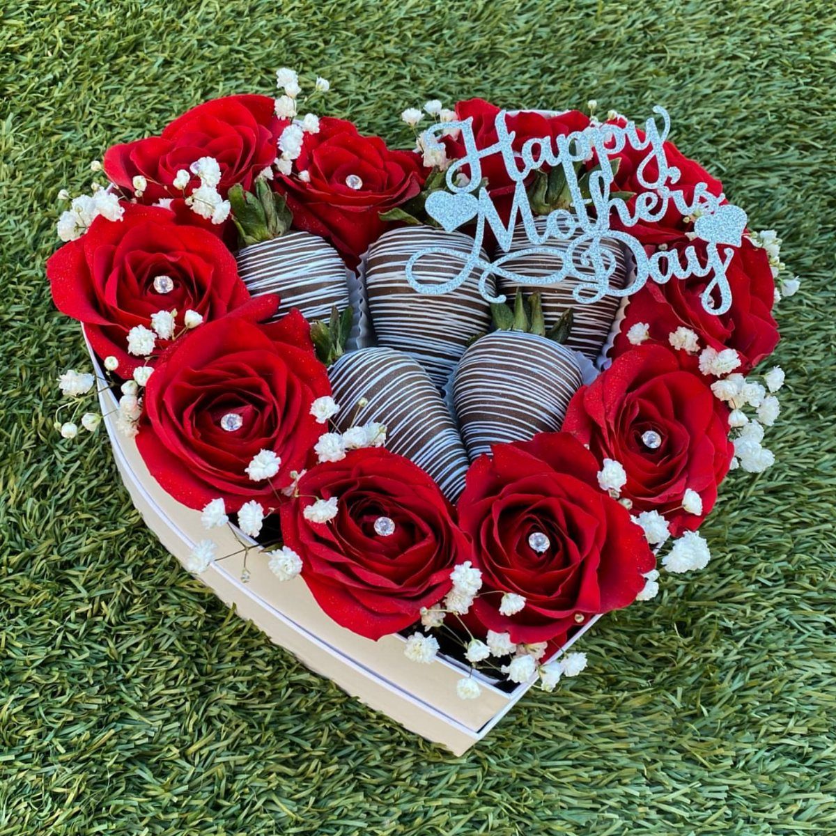 Heart-shaped white box filled with red roses and chocolate strawberries on green grass.