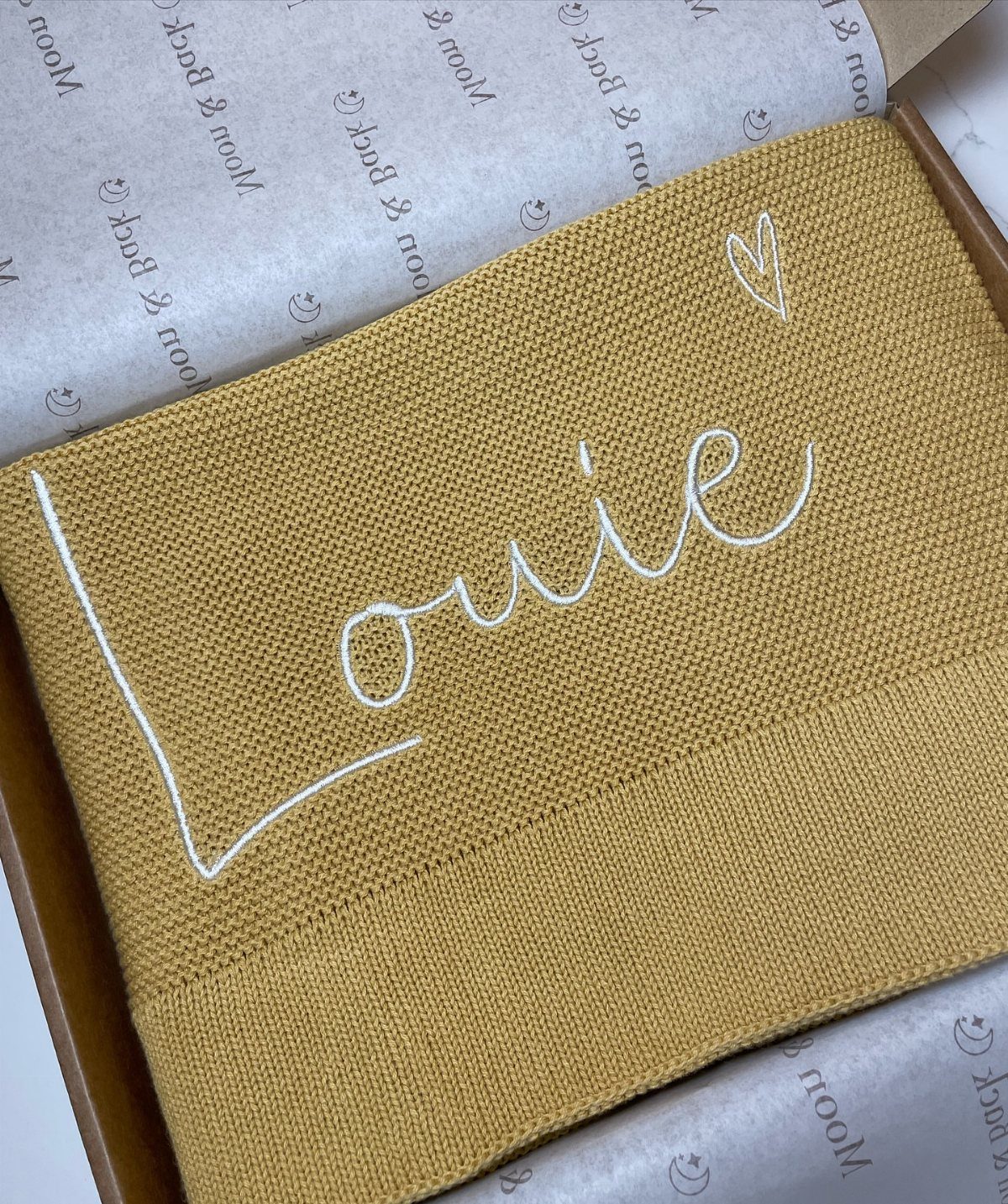Mustard yellow knitted blanket with the name Louie embroidered in white inside a box