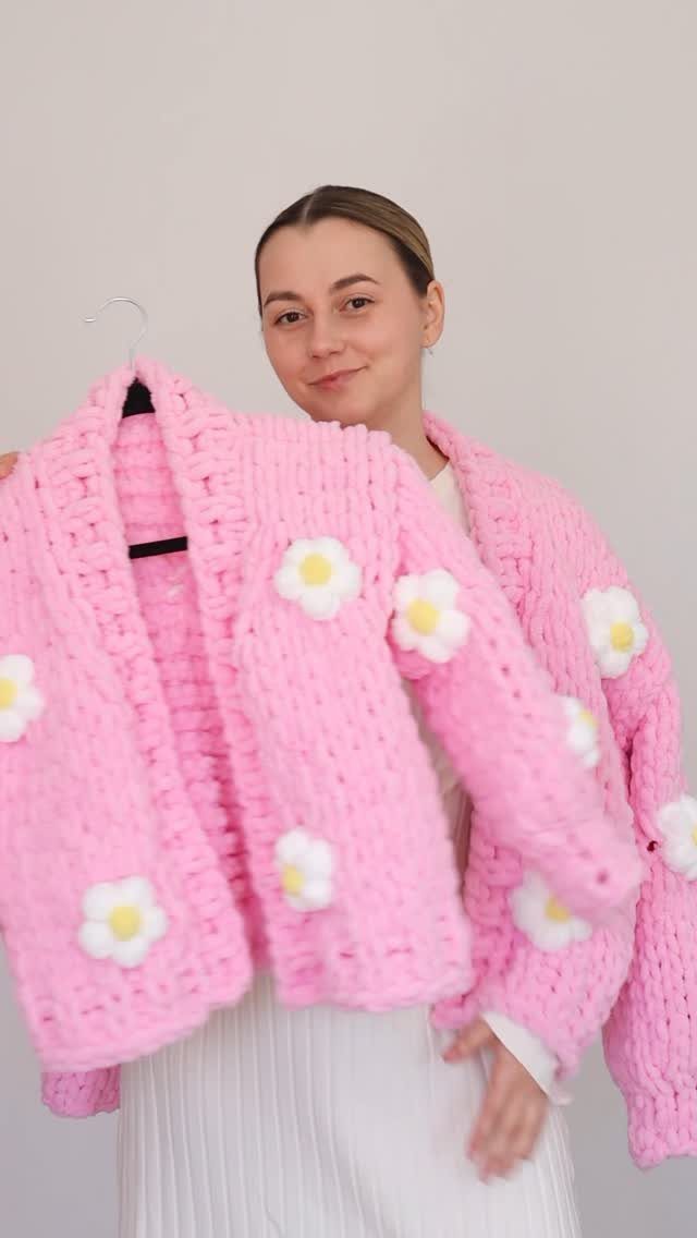 Woman holding a chunky pink knitted cardigan with white daisy flower accents.