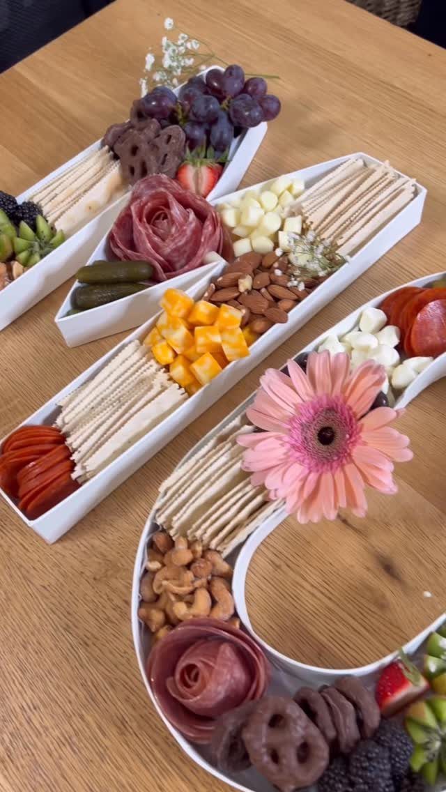 M-shaped charcuterie board with fruits, cheeses, meats, and a pink flower on wood.