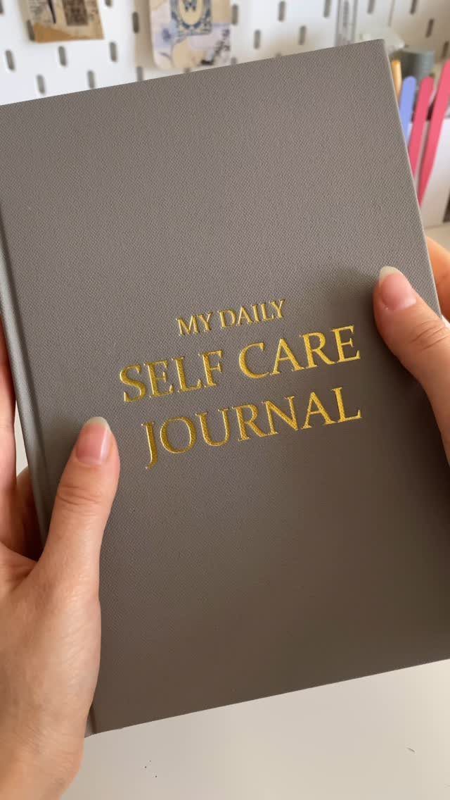 Hands holding a grey hardcover self care journal with gold foil lettering.