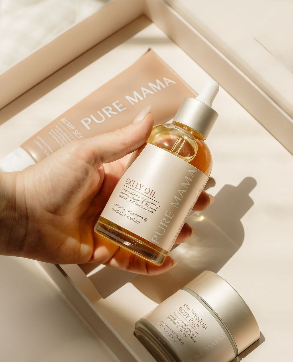 Hand holding a bottle of belly skincare oil on a white tray with creams