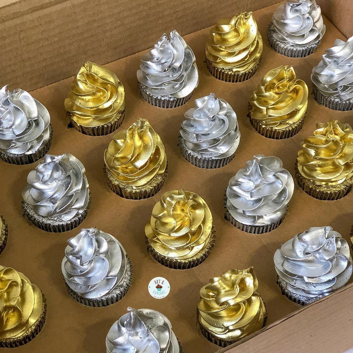 Gold and silver metallic frosted cupcakes arranged in a brown cardboard delivery box.