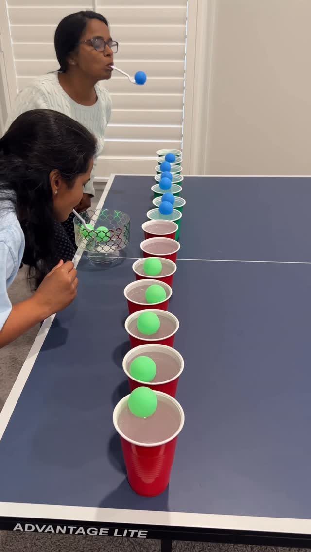 Two women blowing ping pong balls into red cups on a blue table.