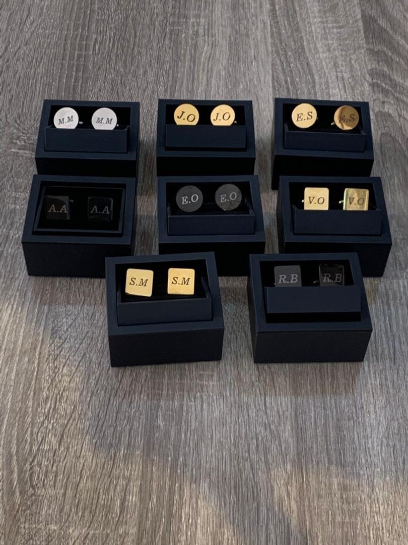 Engraved cufflinks with initials displayed in black velvet boxes on a wooden surface.