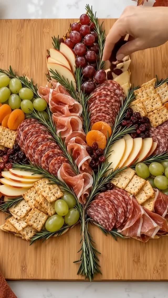 Leaf-shaped charcuterie board with meats, cheeses, grapes, and rosemary on wooden board.