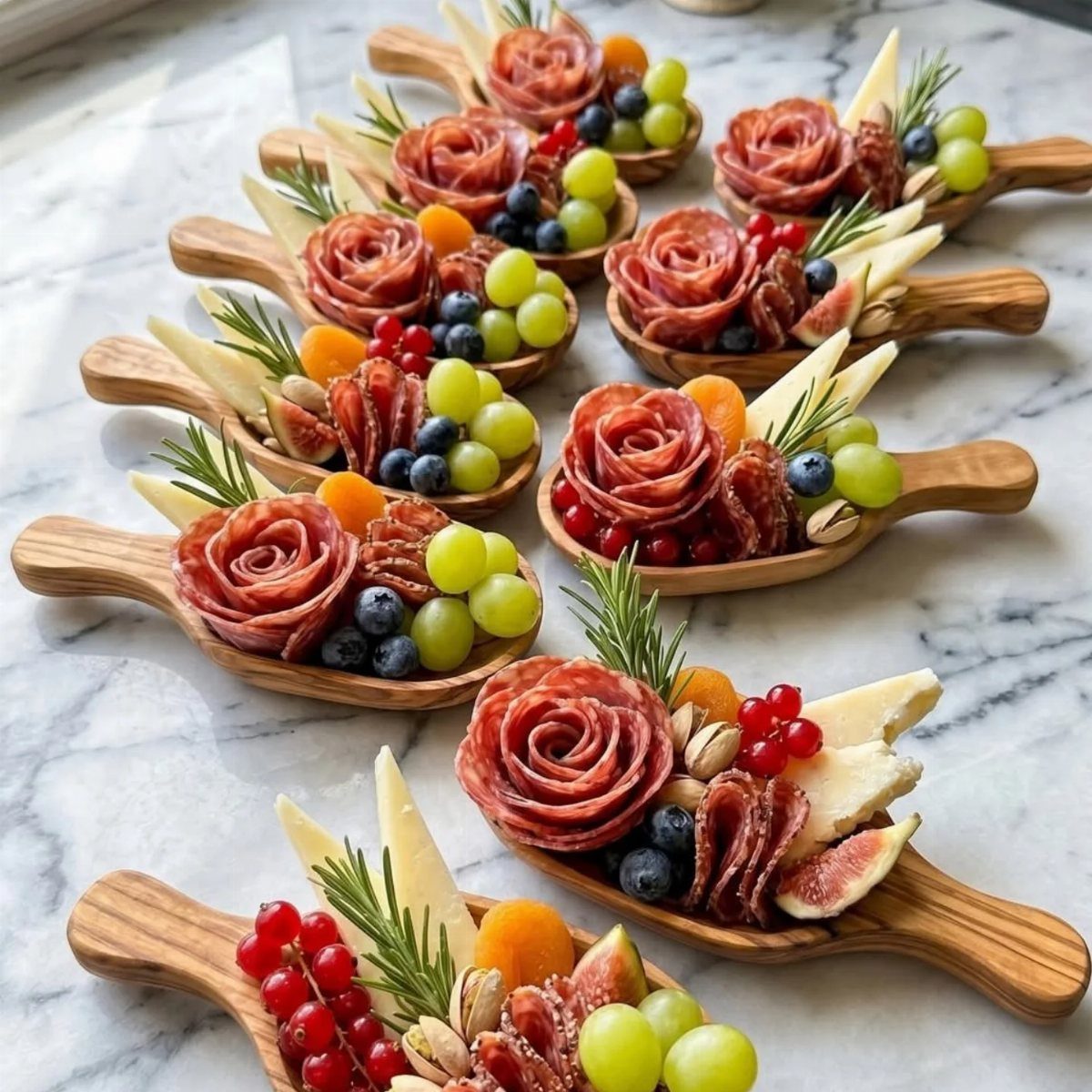 Small wooden spoons filled with salami roses, cheese, fruits, and fresh rosemary sprigs.