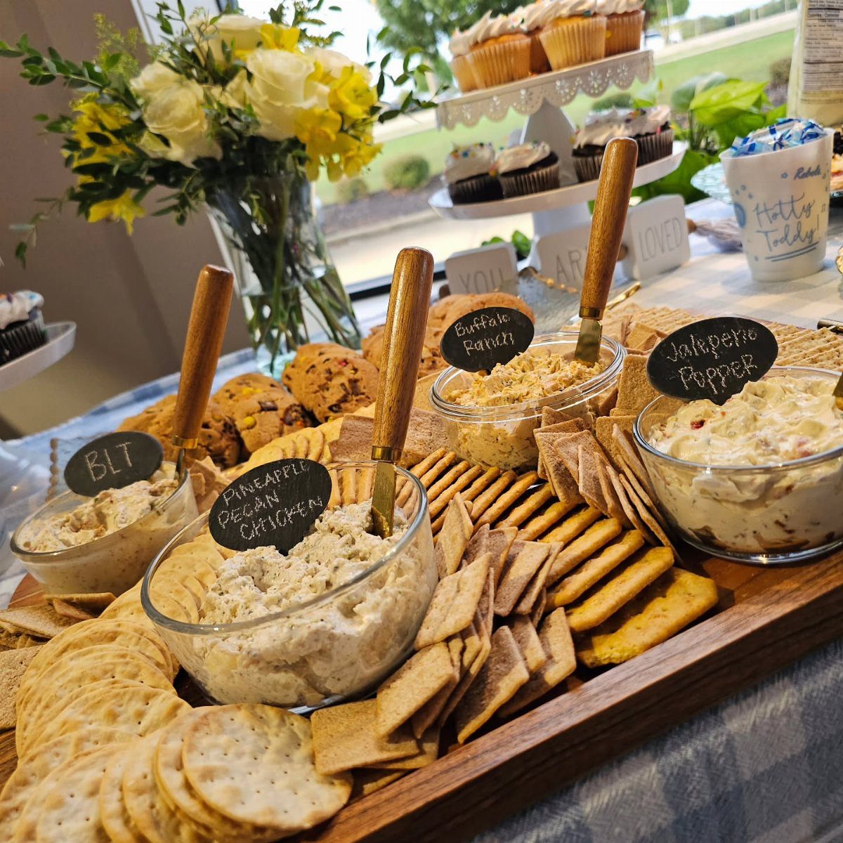 Wooden board with three bowls of creamy dips and assorted crackers and bread.