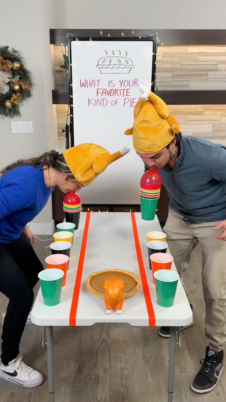 Two adults in turkey hats playing a cup-stacking game at a Thanksgiving party.