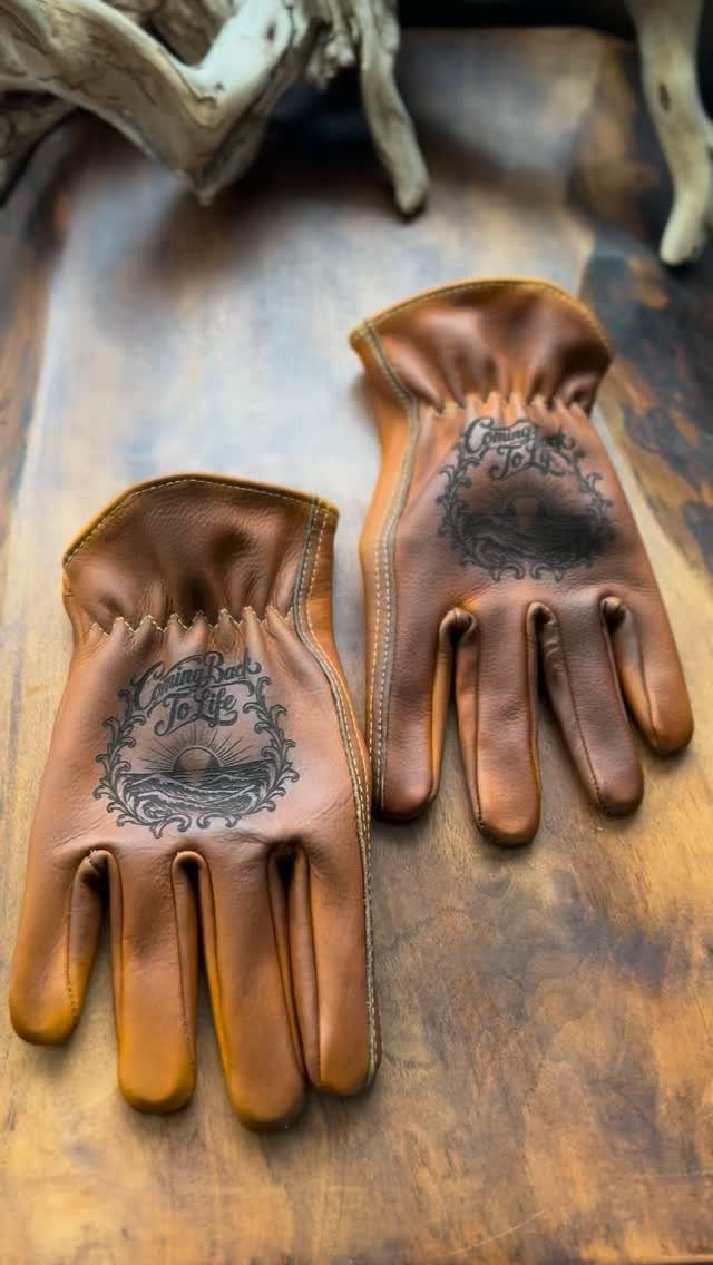 Brown leather gloves with ornate sun and wave illustrations on a wooden surface