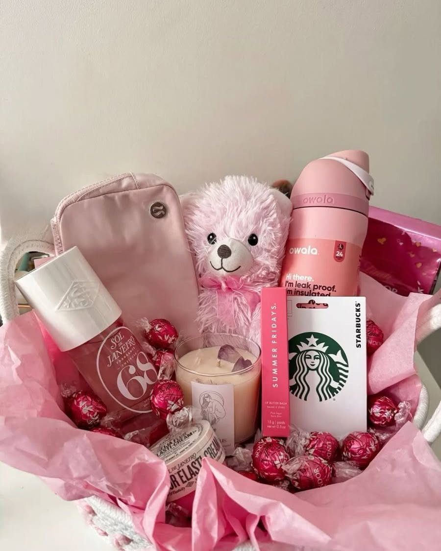 Pink gift basket with teddy bear, water bottle, candle, and chocolates for Valentine's Day.