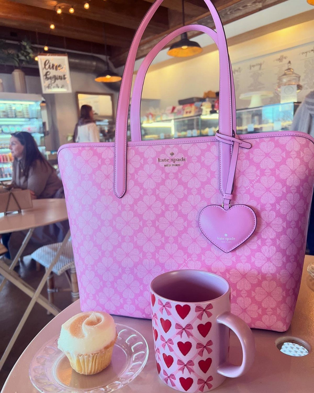 Pink Kate Spade tote bag with heart charm, mug, and cupcake in cafe.