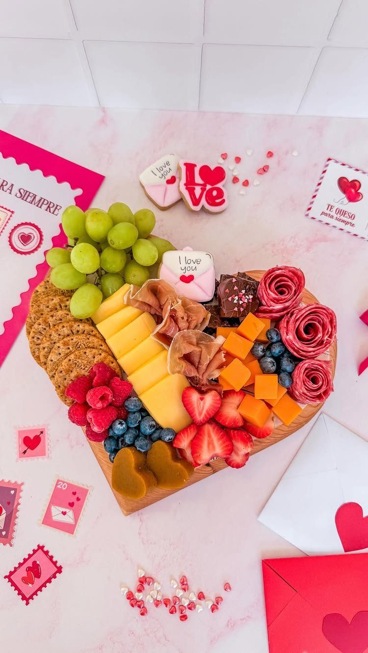 Heart-shaped charcuterie board with fruits, cheeses, and meats on a pink marble background.