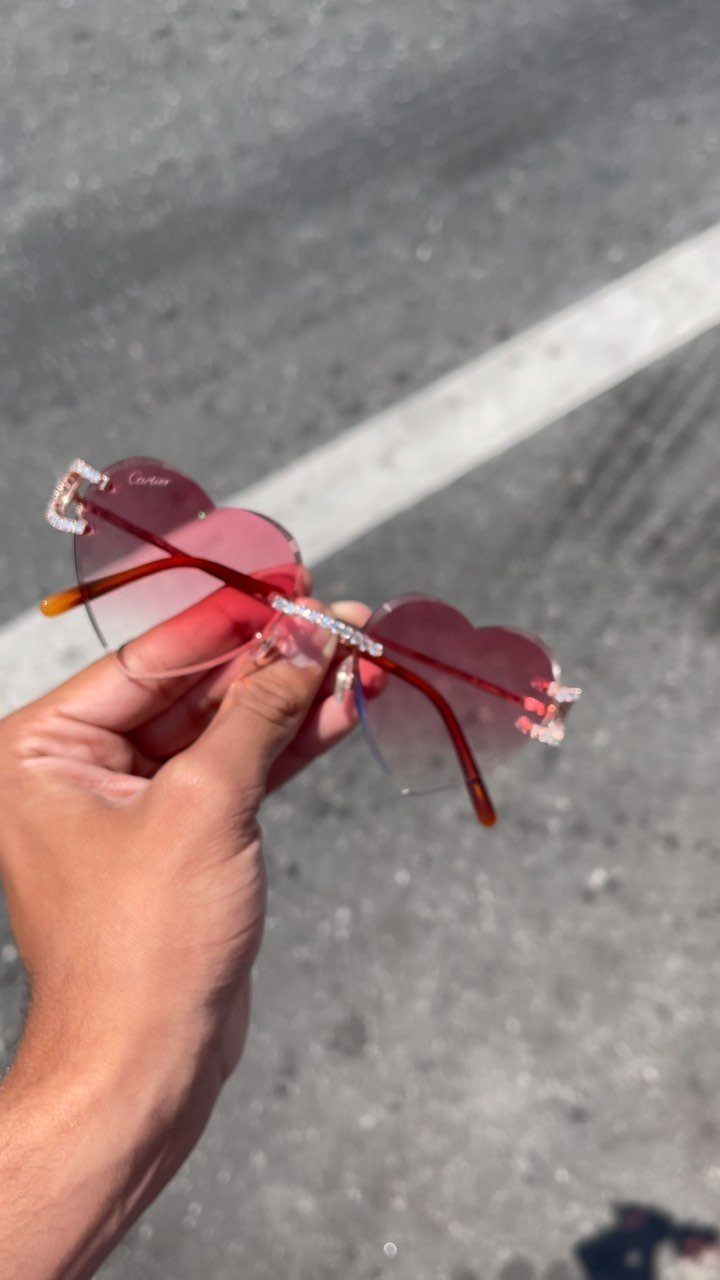 Heart-shaped sunglasses with red lenses and rhinestone frames held by a hand.