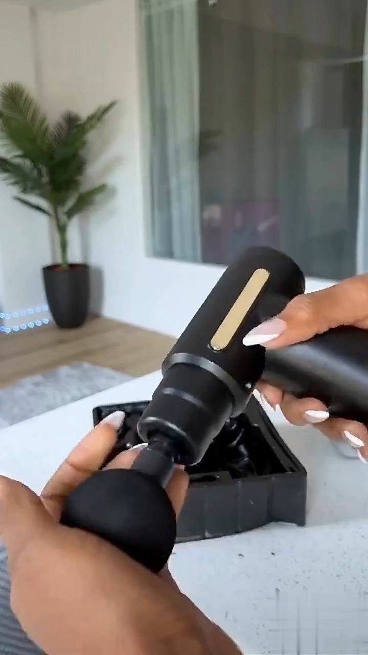 Black handheld massage gun held by hands over its storage case indoors.