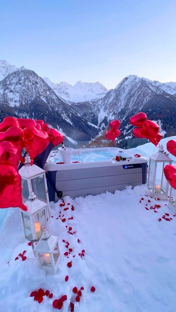 Hot tub in snowy mountains decorated with red heart balloons and rose petals.