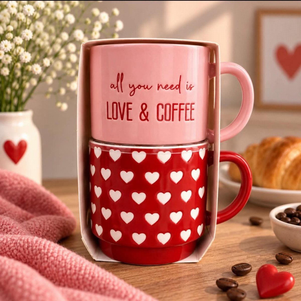 Two heart-themed coffee mugs in a gift set with a croissant and flowers.