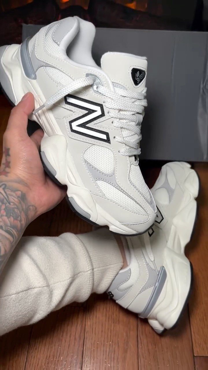 White New Balance sneakers held by a tattooed hand over a wooden floor.