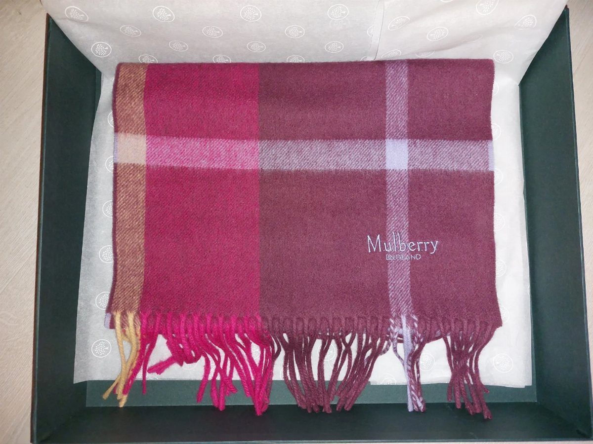 Magenta and purple plaid wool scarf with fringes inside a green gift box.