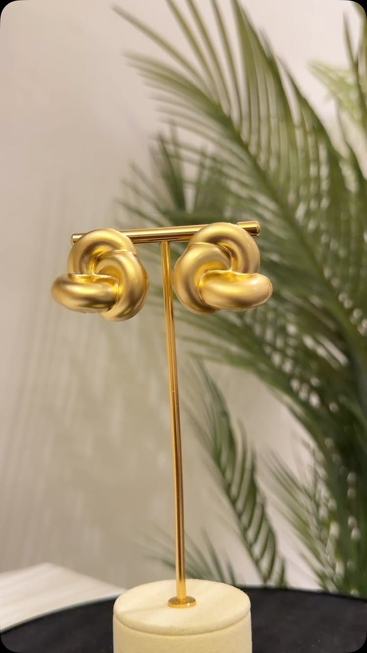 Gold knot-shaped earrings on a gold display stand with a palm leaf background