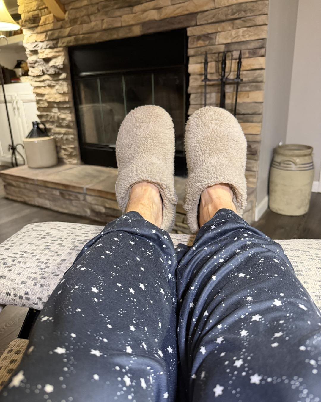 Beige fluffy house slippers and star-patterned pajama pants resting before a stone fireplace.