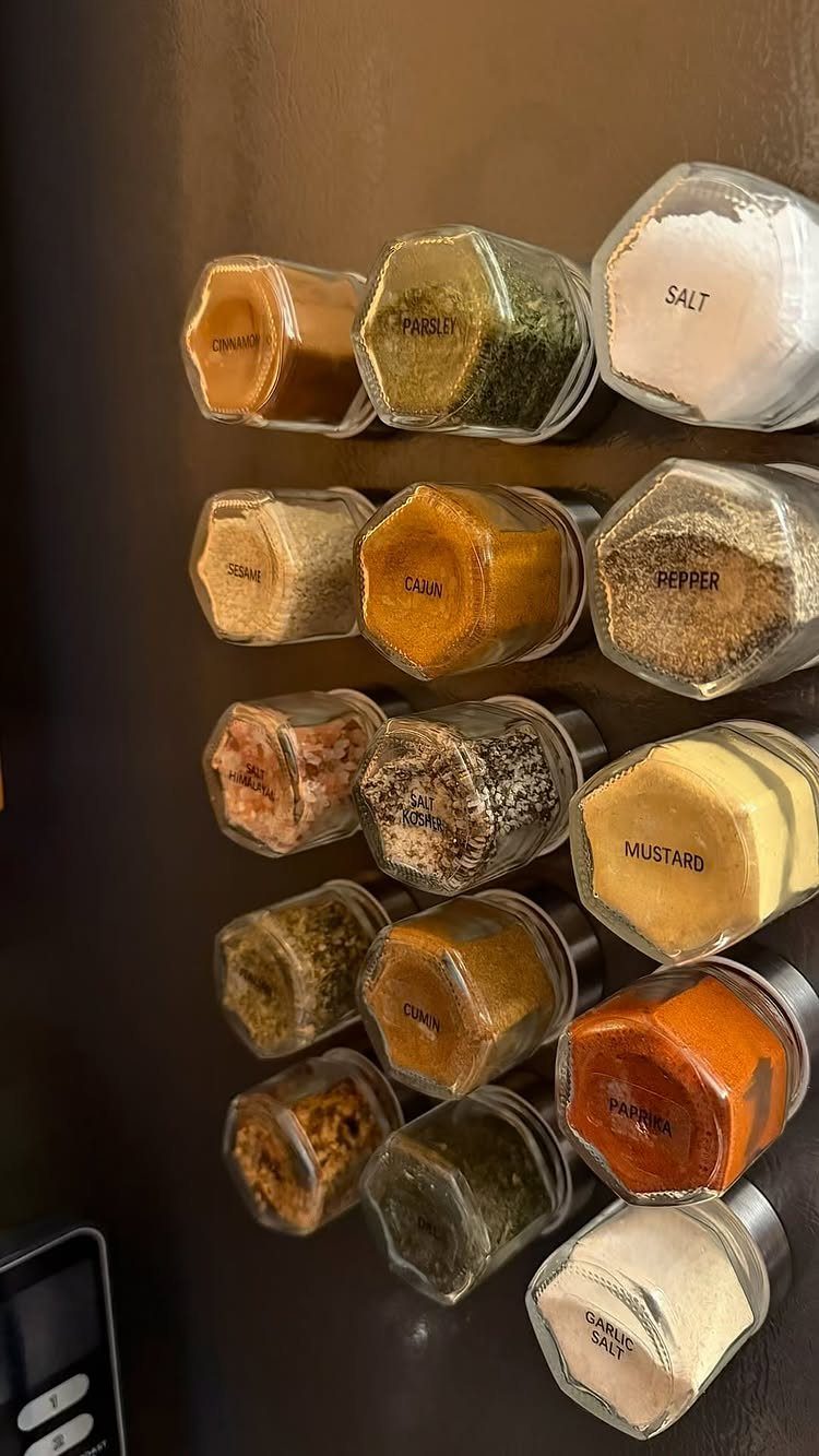 Hexagonal glass spice jars with labels attached magnetically to a dark brown surface.