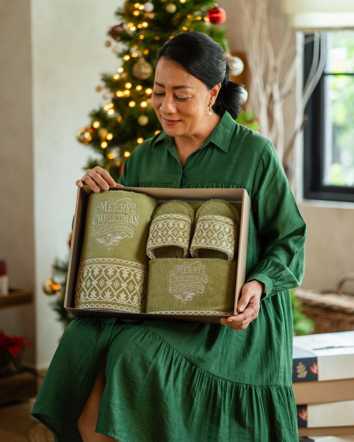 Woman holding a gift box of green Christmas-themed towels near a decorated tree.
